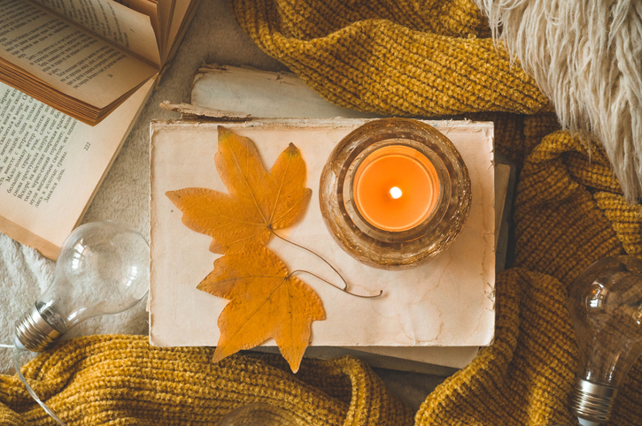 A lit candle and two yellow leaves lying on top of an open book surrounded by other books and a yellow scarf